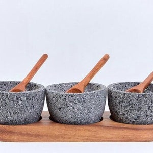 May include: Three small gray stone bowls with wooden spoons, set on a wooden tray. The bowls are made of rough, textured stone and have a rustic look.
