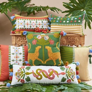 May include: A collection of colourful throw pillows with embroidered designs. The pillows feature a variety of patterns, including geometric shapes, animals, and floral motifs. The pillows are arranged on a wooden shelf with green plants in the background.