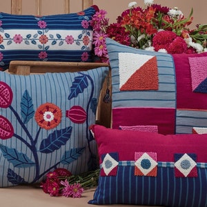 May include: Four decorative throw pillows with embroidered floral and geometric patterns. The pillows are in shades of blue, pink, and white. The pillows are arranged on a brown surface with a bouquet of pink flowers in the foreground.