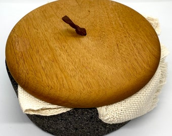 May include: A round, wooden lid with a leather handle sits atop a gray stone mortar and pestle. A white cloth is wrapped around the mortar and pestle.
