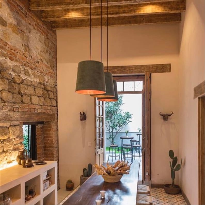 May include: A rustic dining room with a wooden table and chairs. The room has exposed brick walls and wooden beams. There are three pendant lights hanging over the table, each with a dark green shade. The room has a door leading to a patio with plants and furniture.