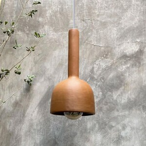 May include: A terracotta pendant light with a bell-shaped shade and a long, cylindrical neck. The light is hanging from a white cord against a textured wall.