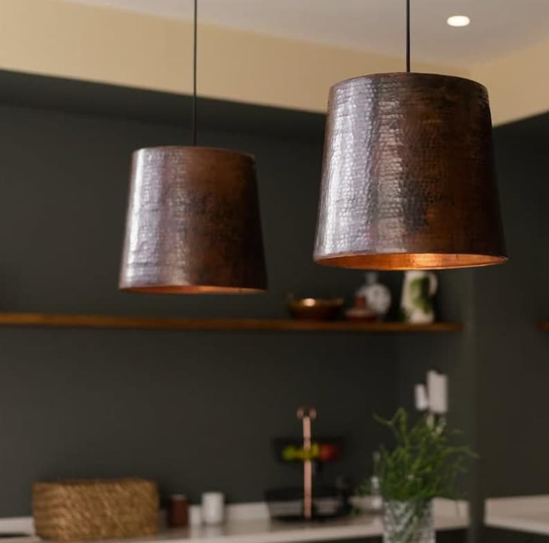 May include: Two copper pendant lights with hammered texture hang from the ceiling in a kitchen setting. The lights are both the same size and shape, and they are both lit.