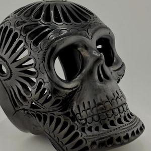 May include: A dark gray, intricately carved skull sculpture. The skull features detailed floral and geometric patterns throughout the surface, with openwork designs creating a lace-like effect. The eye sockets and nasal cavity are open.