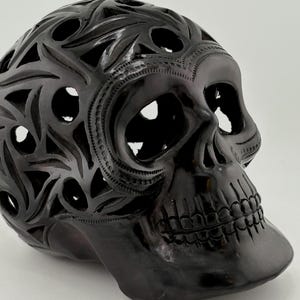 May include: A glossy black decorative skull with intricate cut-out patterns. The skull features detailed carvings around the eye sockets and forehead, and a smooth, polished finish. The design is a unique piece of home decor.