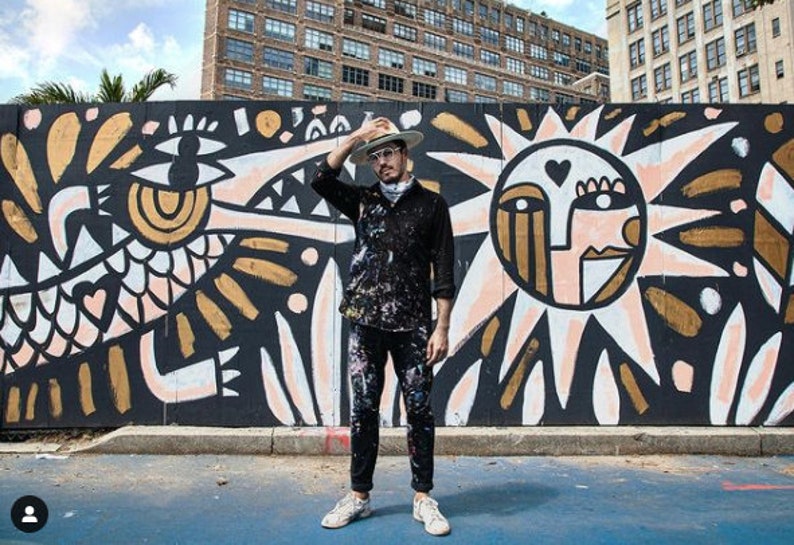 May include: A person wearing a black shirt and a hat stands in front of a large mural. The mural features a black, white, and gold design with a sun and a bird-like creature.