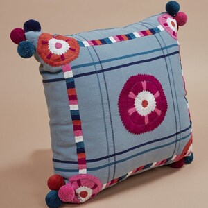 May include: A blue throw pillow with a checkered pattern and embroidered flowers in shades of pink, red, and white. The pillow has pom-poms on the corners.
