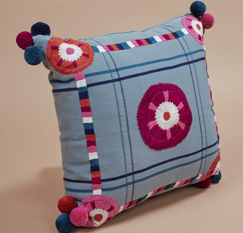 May include: A blue throw pillow with a checkered pattern and embroidered flowers in shades of pink, red, and white. The pillow has pom-poms on the corners.