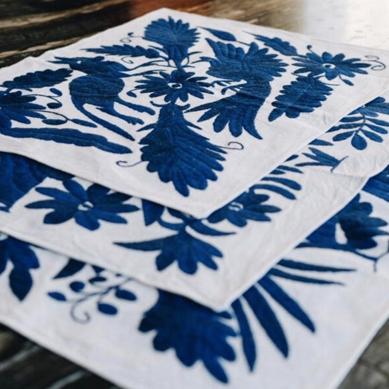 May include: Three white linen placemats with blue embroidered floral and bird designs. The placemats are stacked on top of each other.
