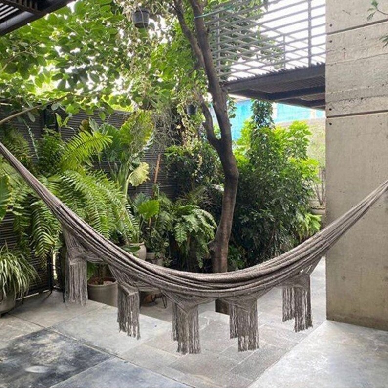 Puede incluir: Una hamaca gris con flecos cuelga entre dos &aacute;rboles en un exuberante jard&iacute;n verde. La hamaca est&aacute; hecha de tejido tejido y tiene un estilo relajado y bohemio.