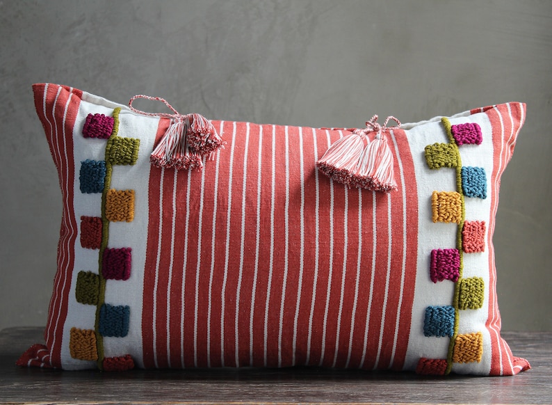May include: Red and white striped lumbar pillow with colorful embroidered squares and tassels on each end.