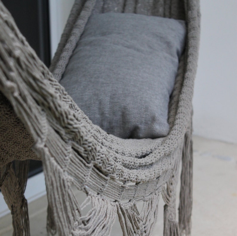 Puede incluir: Una hamaca gris con detalles de flecos. La hamaca est&aacute; hecha de un material tejido y tiene una almohada gris en su interior.