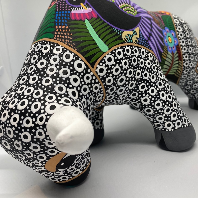May include: Black and white ceramic bull figurine with a white nose and a colorful floral design on the back. The bull is painted with a black and white polka dot pattern.