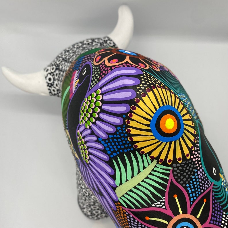 May include: A black ceramic bull figurine with colorful painted details, including flowers, leaves, and birds. The bull has white horns and a white spot on its back.