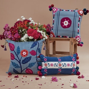 May include: Three decorative throw pillows with blue and white striped fabric and colorful floral embroidery. The pillows have pom-poms in shades of pink and red. The pillows are arranged on a wooden stool with a bouquet of pink and white flowers in front.
