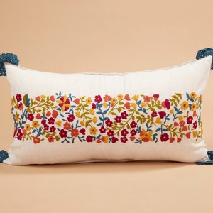May include: A white rectangular pillow with a floral embroidery design in shades of red, yellow, and blue. The pillow has blue tassels on each end.