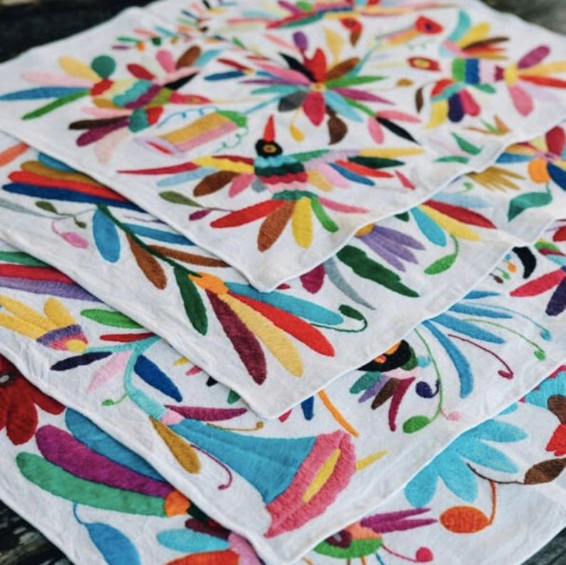 May include: A stack of white fabric napkins with colorful embroidered floral designs. The designs feature flowers, leaves, and birds in shades of pink, blue, yellow, green, and red.