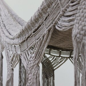 Puede incluir: Una hamaca de macram&eacute; beige con un patr&oacute;n tejido y detalles de flecos. La hamaca est&aacute; colgada del techo y est&aacute; hecha de fibras naturales.