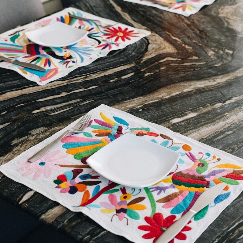 May include: Two white plates with silver forks on top of colorful embroidered placemats. The placemats have a floral design with birds and flowers. The plates are on a dark brown table.
