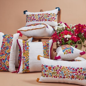 May include: A set of six white embroidered throw pillows with floral patterns in shades of pink, red, yellow, and blue. The pillows have tassels and are arranged on a light brown surface.