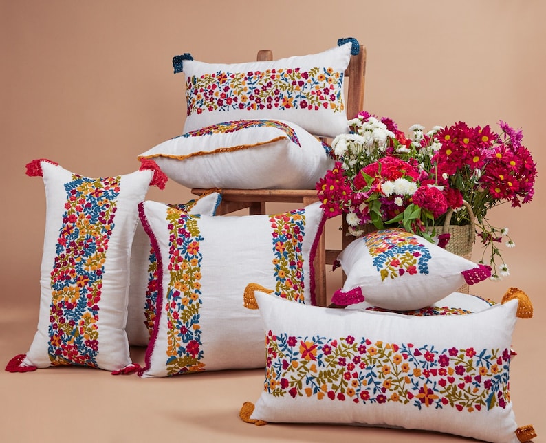 May include: A set of six white embroidered throw pillows with floral patterns in shades of pink, red, yellow, and blue. The pillows have tassels and are arranged on a light brown surface.