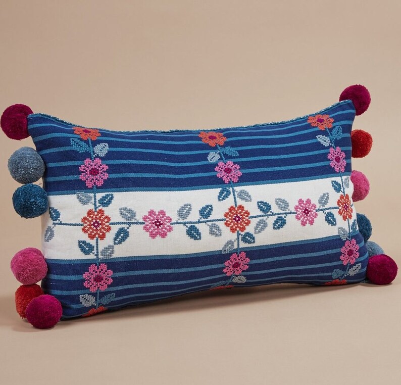May include: A blue and white striped pillow with embroidered pink flowers and green leaves. The pillow has a pom-pom trim in pink and blue.
