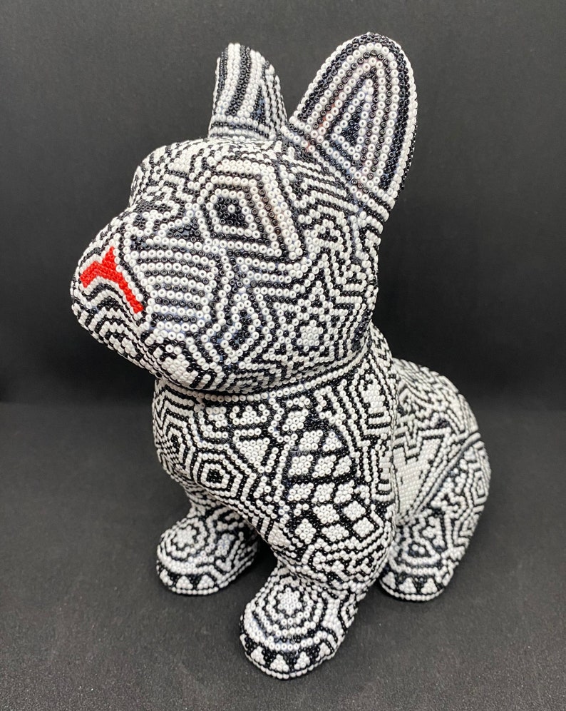 May include: A black and white beaded figurine of a French bulldog. The figurine is made of small, round beads that are arranged in a geometric pattern. The bulldog has a red nose and is sitting on its hind legs.