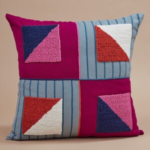 May include: A square throw pillow with a patchwork design. The pillow is made of four different fabrics, each with a different color and pattern. The fabric is a mix of pink, blue, red, and white. The pillow has a geometric design with triangles and squares.