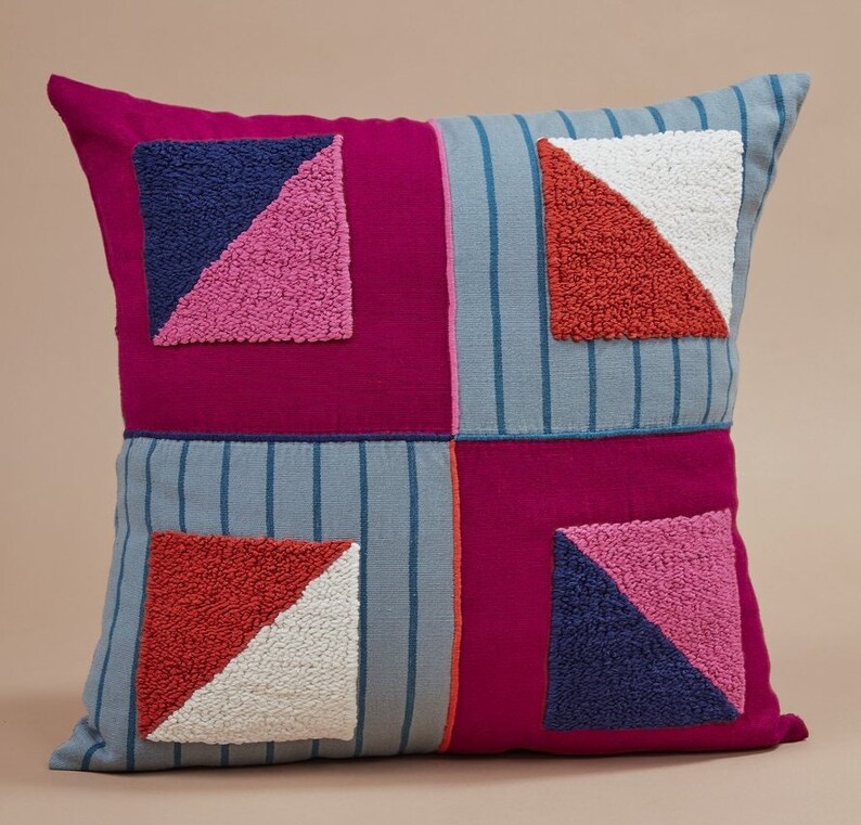 May include: A square throw pillow with a patchwork design. The pillow is made of four different fabrics, each with a different color and pattern. The fabric is a mix of pink, blue, red, and white. The pillow has a geometric design with triangles and squares.