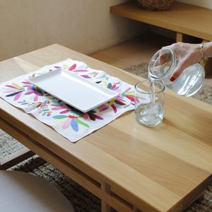 May include: A white rectangular plate sits on a colorful embroidered table runner on a light wood table. A glass is being filled with water from a clear glass pitcher.