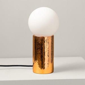 May include: A modern table lamp featuring a white globe-shaped shade atop a textured copper-colored cylindrical base. The lamp has a hammered metal finish and a black power cord. The lamp is designed for indoor use.