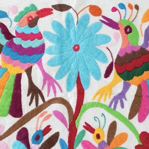 May include: A white fabric with colorful embroidery of two birds with long, multi-colored tails, a large blue flower, and smaller flowers. The birds are surrounded by green leaves and stems.