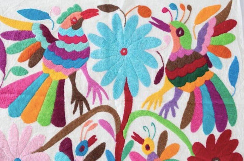 May include: A white fabric with colorful embroidery of two birds with long, multi-colored tails, a large blue flower, and smaller flowers. The birds are surrounded by green leaves and stems.