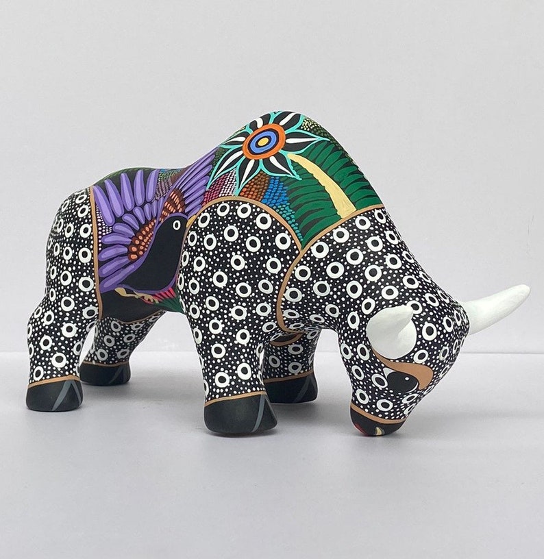 May include: A black and white ceramic bull figurine with colorful painted details, including a purple bird with a black head and a green and yellow flower on the back.
