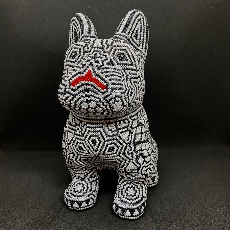 May include: A black and white beaded figurine of a dog. The dog is sitting with its legs crossed and has a red nose.