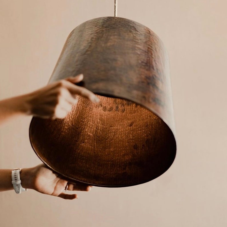 May include: A hammered copper pendant light fixture with a warm, aged finish. The light is suspended from a cord and has a wide, bell-shaped shade.