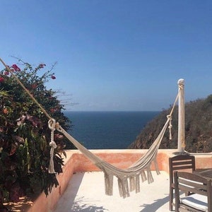 Puede incluir: Una hamaca blanca cuelga entre dos postes de madera en un patio con vistas al oc&eacute;ano. La hamaca est&aacute; hecha de tejido tejido con borlas en los extremos. El oc&eacute;ano es azul y el cielo est&aacute; despejado.