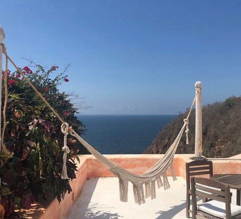 Puede incluir: Una hamaca blanca cuelga entre dos postes de madera en un patio con vistas al oc&eacute;ano. La hamaca est&aacute; hecha de tejido tejido con borlas en los extremos. El oc&eacute;ano es azul y el cielo est&aacute; despejado.