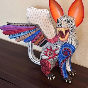 May include: A hand-painted alebrije sculpture of a winged creature with large orange ears, sharp teeth, and a long tongue. The body is covered in intricate black and white patterns, accented with vibrant red, blue, and purple geometric designs.