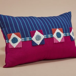 May include: A pink and blue striped throw pillow with colorful embroidered geometric designs. The pillow has a blue and white striped fabric on the front and a solid pink fabric on the back.