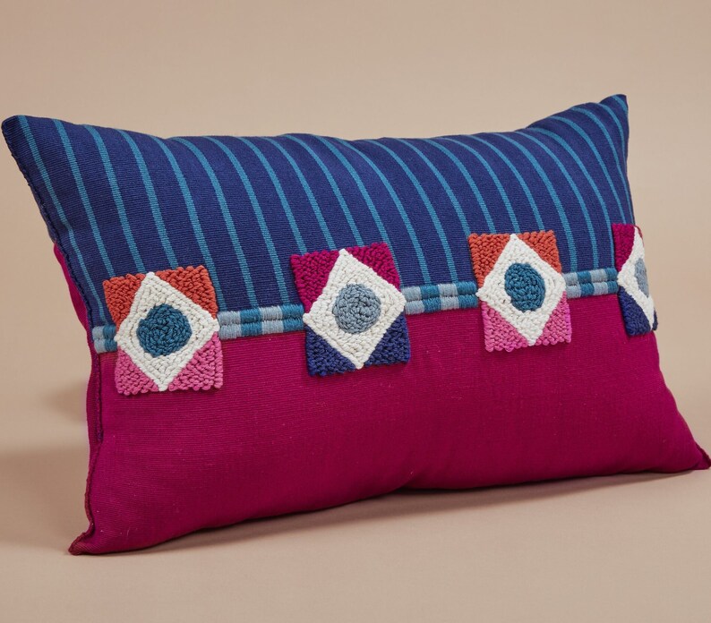 May include: A pink and blue striped throw pillow with colorful embroidered geometric designs. The pillow has a blue and white striped fabric on the front and a solid pink fabric on the back.
