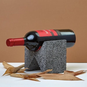 May include: A gray stone wine bottle holder with a bottle of red wine resting in it. The holder is made of rough, textured stone and has a modern, minimalist design.