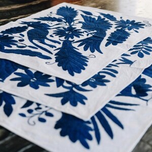 May include: Three white cotton placemats with blue embroidered floral designs. The designs feature stylized flowers, leaves, and birds.