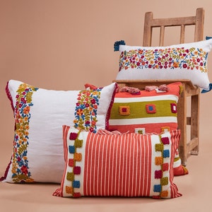 May include: Four decorative throw pillows with embroidered floral patterns and colorful stripes. The pillows are arranged on a wooden chair with a light brown background.