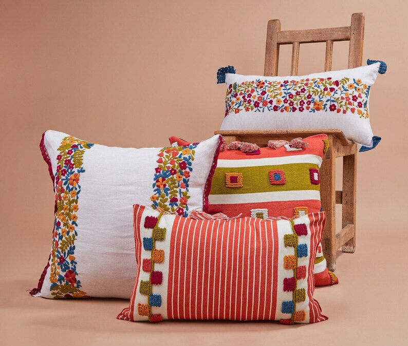 May include: Four decorative throw pillows with embroidered floral patterns and colorful stripes. The pillows are arranged on a wooden chair with a light brown background.