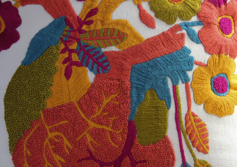 May include: A white pillow with colorful embroidered flowers and leaves. The flowers are yellow, orange, pink, and red. The leaves are green and yellow. The embroidery is done in a variety of stitches, including satin stitch, French knots, and seed stitch.