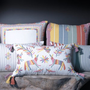 May include: Five colourful embroidered throw pillows with tassels. The pillows are arranged in a row on a wooden surface. The pillows feature various patterns and designs, including stripes, geometric shapes, and floral motifs.