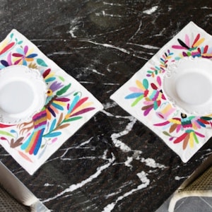 May include: Two white plates with scalloped edges are placed on top of colorful embroidered placemats. The placemats feature a vibrant floral and bird design with a white background.