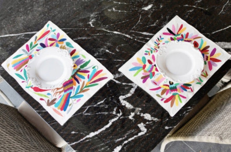May include: Two white plates with scalloped edges are placed on top of colorful embroidered placemats. The placemats feature a vibrant floral and bird design with a white background.
