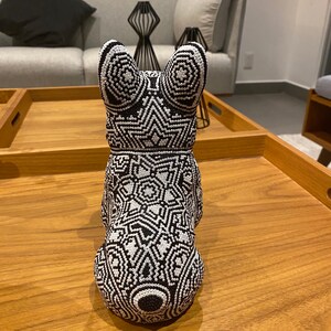 May include: A black and white beaded figurine of a dog. The figurine is sitting on a wooden surface. The dog is wearing a geometric pattern.
